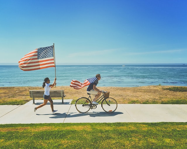 A woman sprinting alongside a man on a bike on a seaside bike lane. Both are carrying American flags.